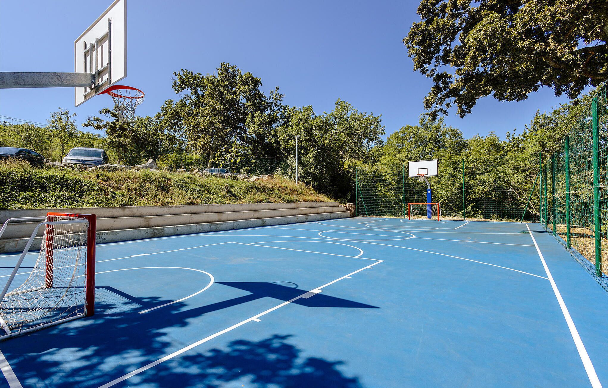 An outdoor basketball court with a blue surface, a basketball hoop, and a goal post.