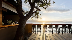 Una terraza con sillas que da a un cuerpo de agua al atardecer, un árbol y un edificio con ventana y plantas en macetas.