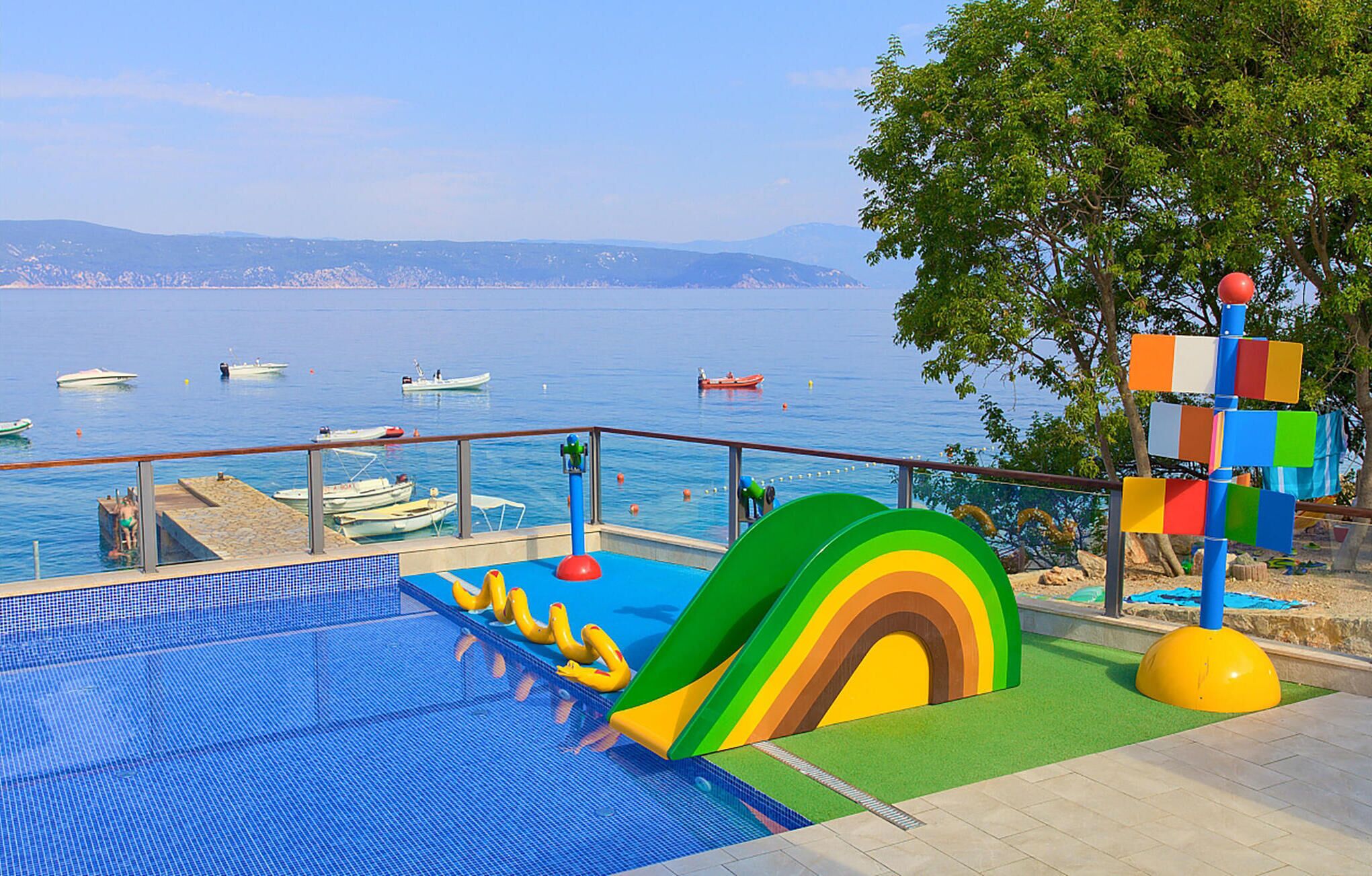 Ein Poolbereich mit Rutsche, Blick auf das Meer und Booten im Hintergrund.