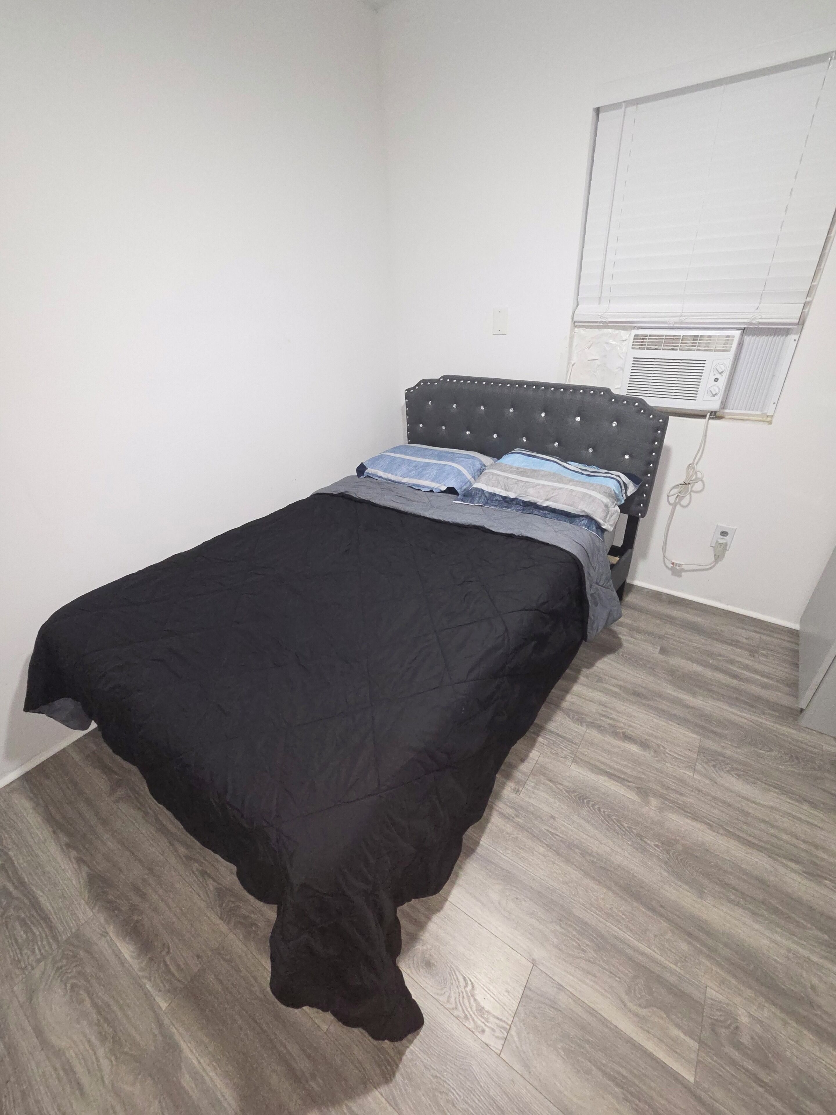 A room with a bed, a window with blinds, and an air conditioning unit.