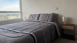 Un dormitorio con una cama, una mesita de noche, una lámpara y vistas a la ciudad a través de una ventana.