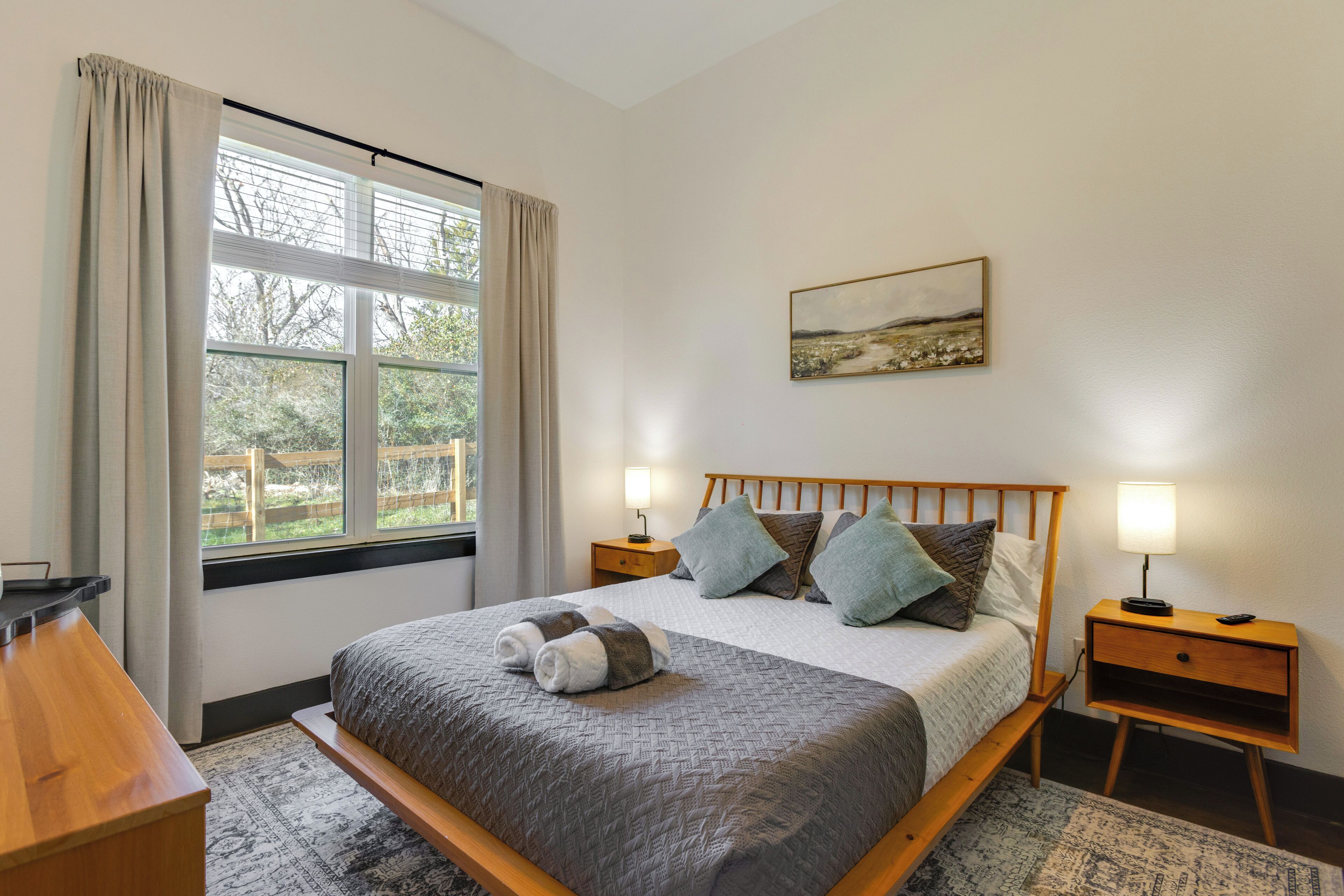 Une chambre à coucher bien rangée, avec un lit, des tables de chevet, une fenêtre avec des rideaux et un tableau accroché au mur.