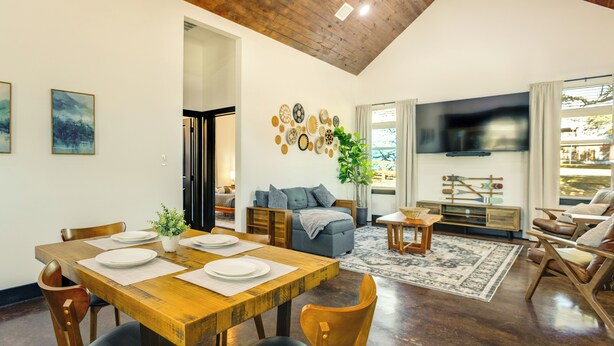 Een ruime woonkamer met een houten eettafel voor vier personen, een comfortabele bank, een flatscreen televisie en een groot raam met gordijnen.
