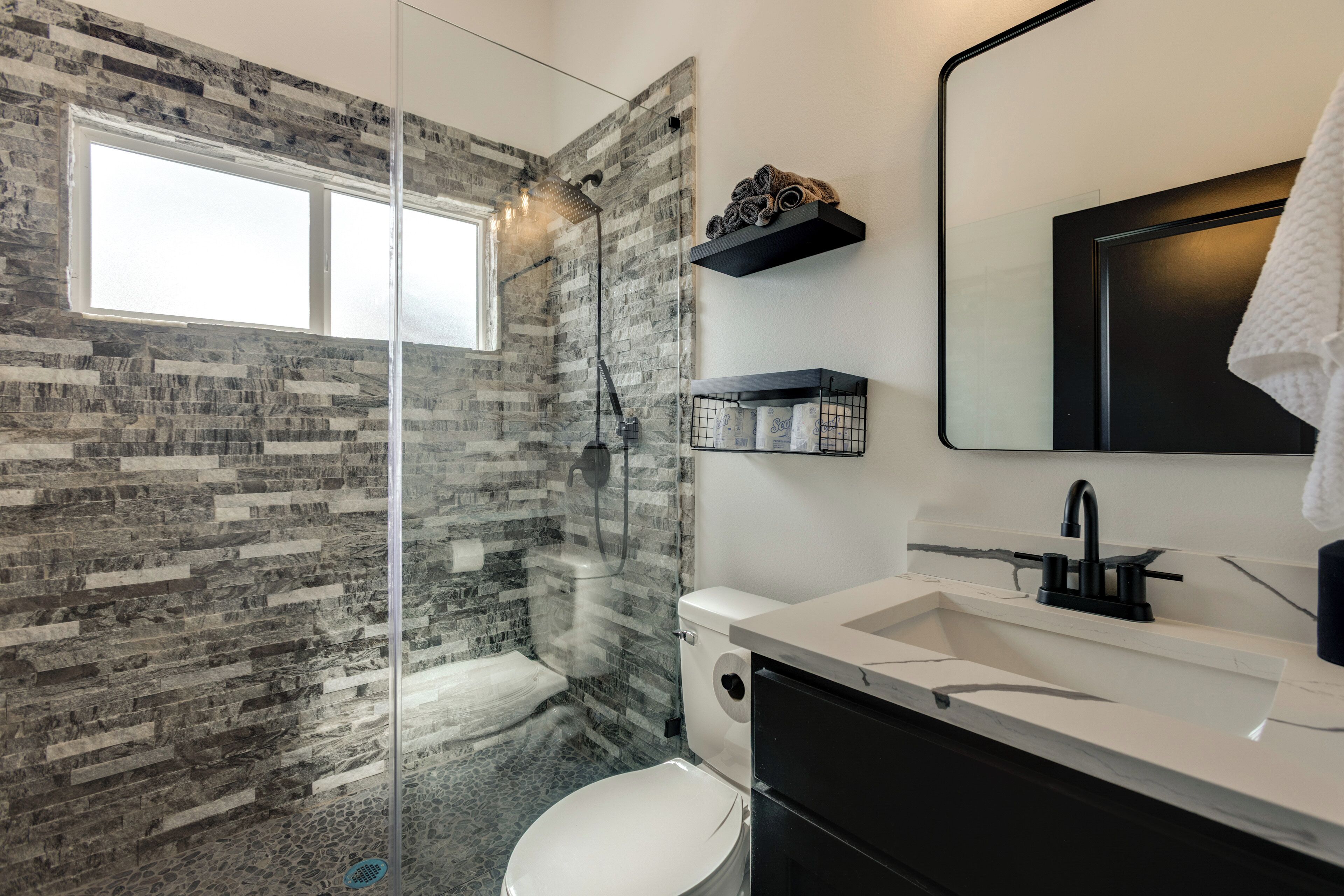 A modern bathroom with a glass-enclosed shower, a white toilet, a black sink with a marble countertop, and a large mirror.