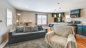 Un salon moderne avec un canapé gris, un fauteuil beige et un coin repas comprenant une table et des chaises. La cuisine est dotée de meubles bleu foncé et d’un four à micro-ondes noir.