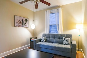 Ein Wohnzimmer mit grauem Sofa, zwei gemusterten Kissen, Deckenventilator, Stehlampe, Beistelltisch und einer gerahmten Pflanze an der Wand.