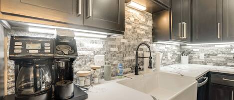 A modern kitchen with dark cabinets, a white countertop, a coffee maker, and a stone backsplash.