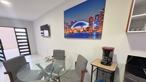 Una sala de estar moderna con una mesa de comedor de vidrio rodeada de sillas tapizadas en gris. Una cafetera y un dispensador de agua están sobre una pequeña mesa auxiliar de madera. Un gran cuadro de un paisaje urbano decora la pared.