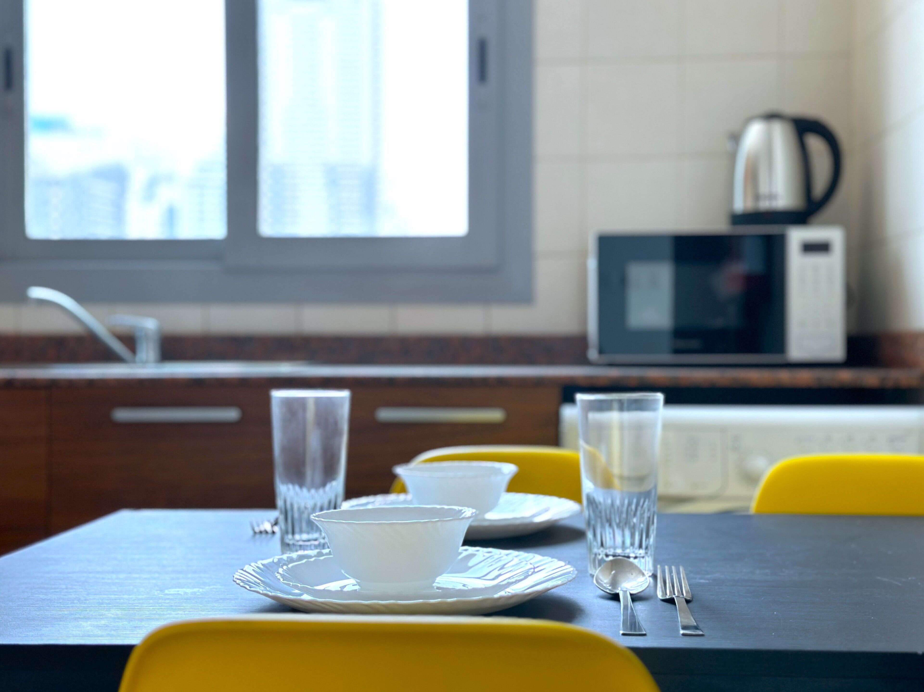 Een eettafel met een witte kom, een glas en bestek, voor een gele stoel.