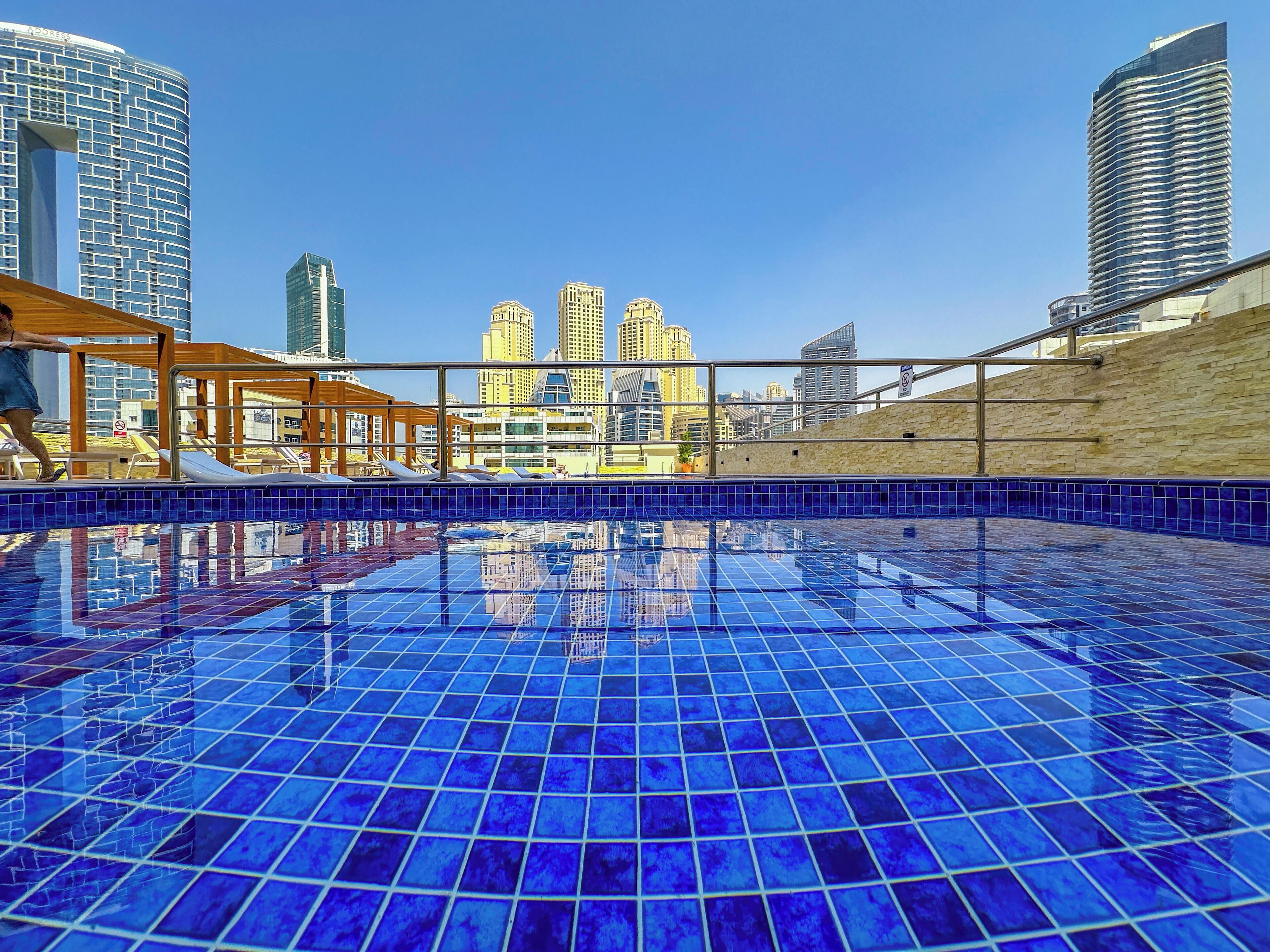 A rooftop pool with blue tiles, surrounded by city buildings and a wooden pergola.