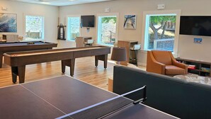 A pool table, ping pong table, and billiards equipment in a well-lit room with large windows and a view of the outdoors.