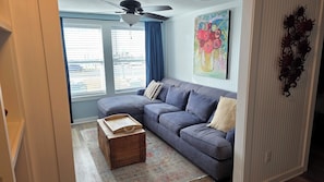 A living room with a grey sectional sofa, a wooden chest of drawers, a patterned rug, a ceiling fan, and a window with blinds. - Sunsets - Downtown Duck + Indoor P00L and Beach access (Kitty Hawk)