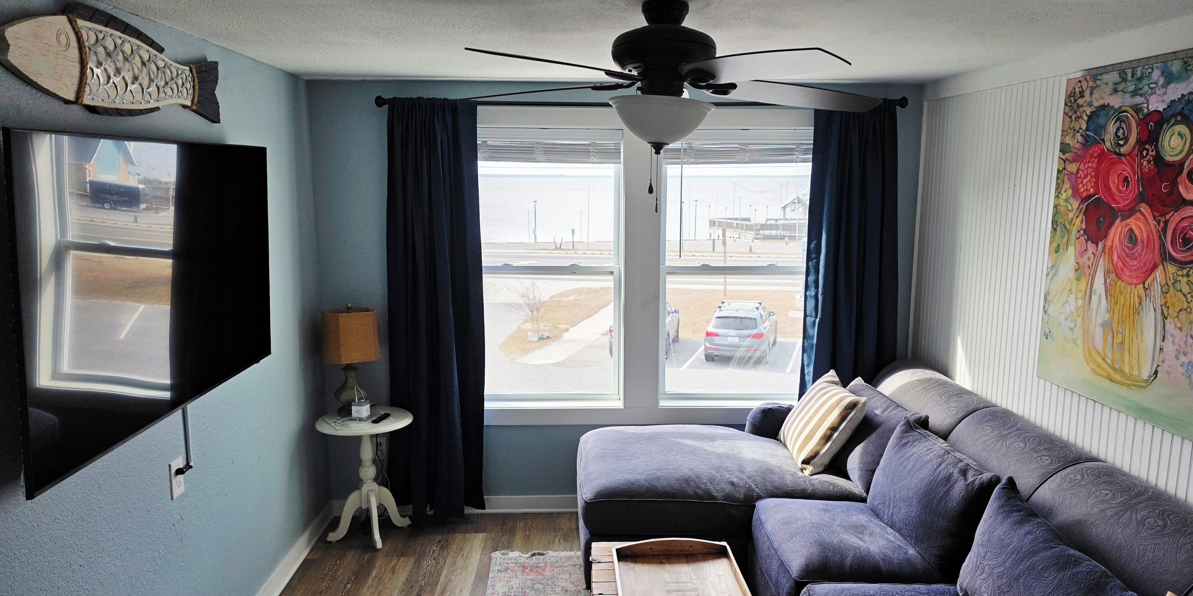 A living room with a dark blue sofa, a ceiling fan, a flat-screen TV mounted on the wall, a small side table with a lamp, and a window with curtains.