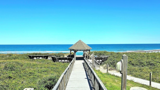 A wooden boardwalk leading to a gazebo with benches, overlooking a beach and ocean.