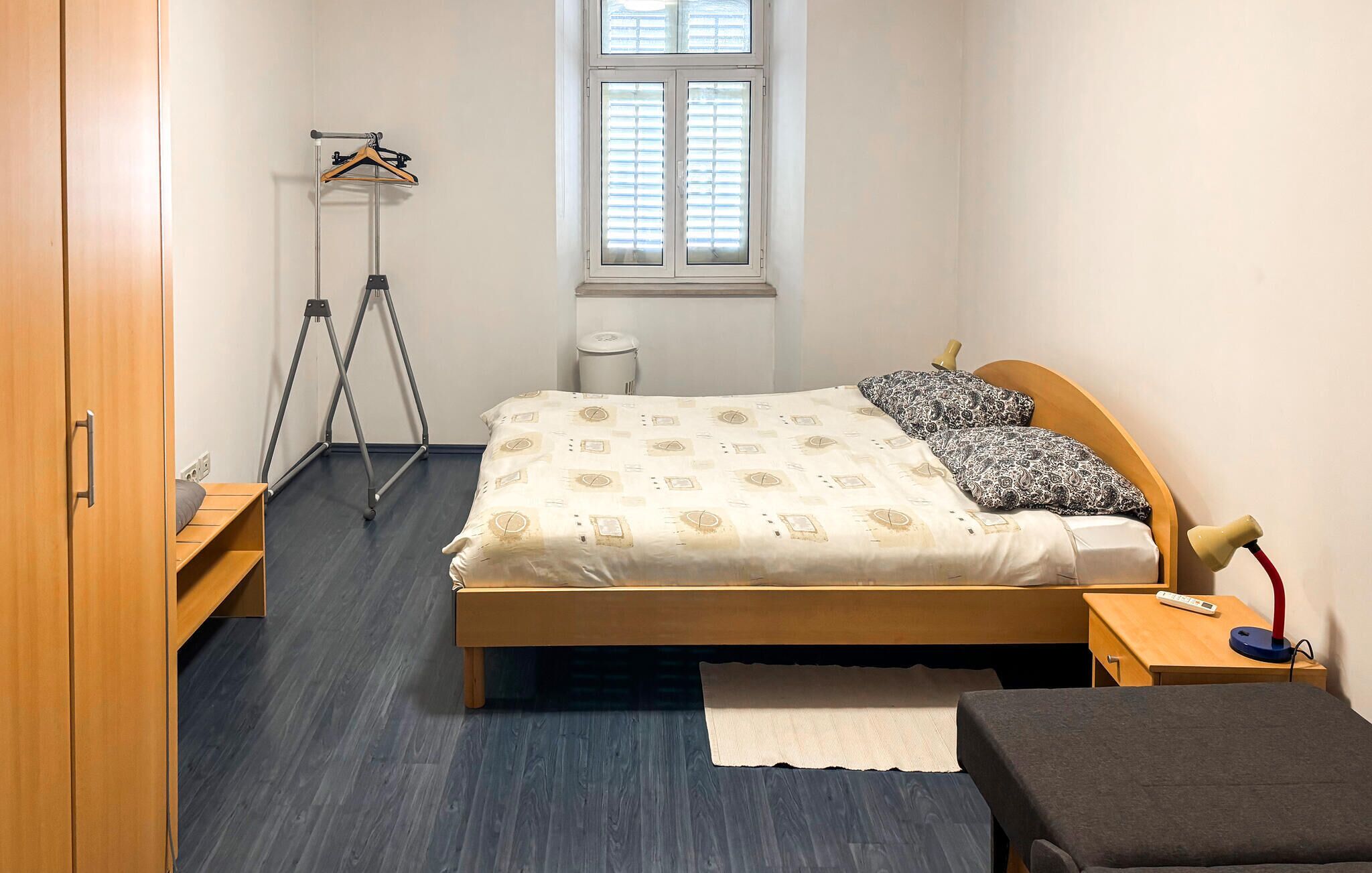 A small, neatly organized bedroom with a bed, bedside tables, a wardrobe, a tripod, and a window.