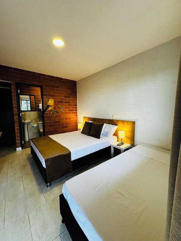 Une chambre d’hôtel avec deux lits, un mur décoratif en briques, une petite salle de bain et une table de chevet avec une lampe.