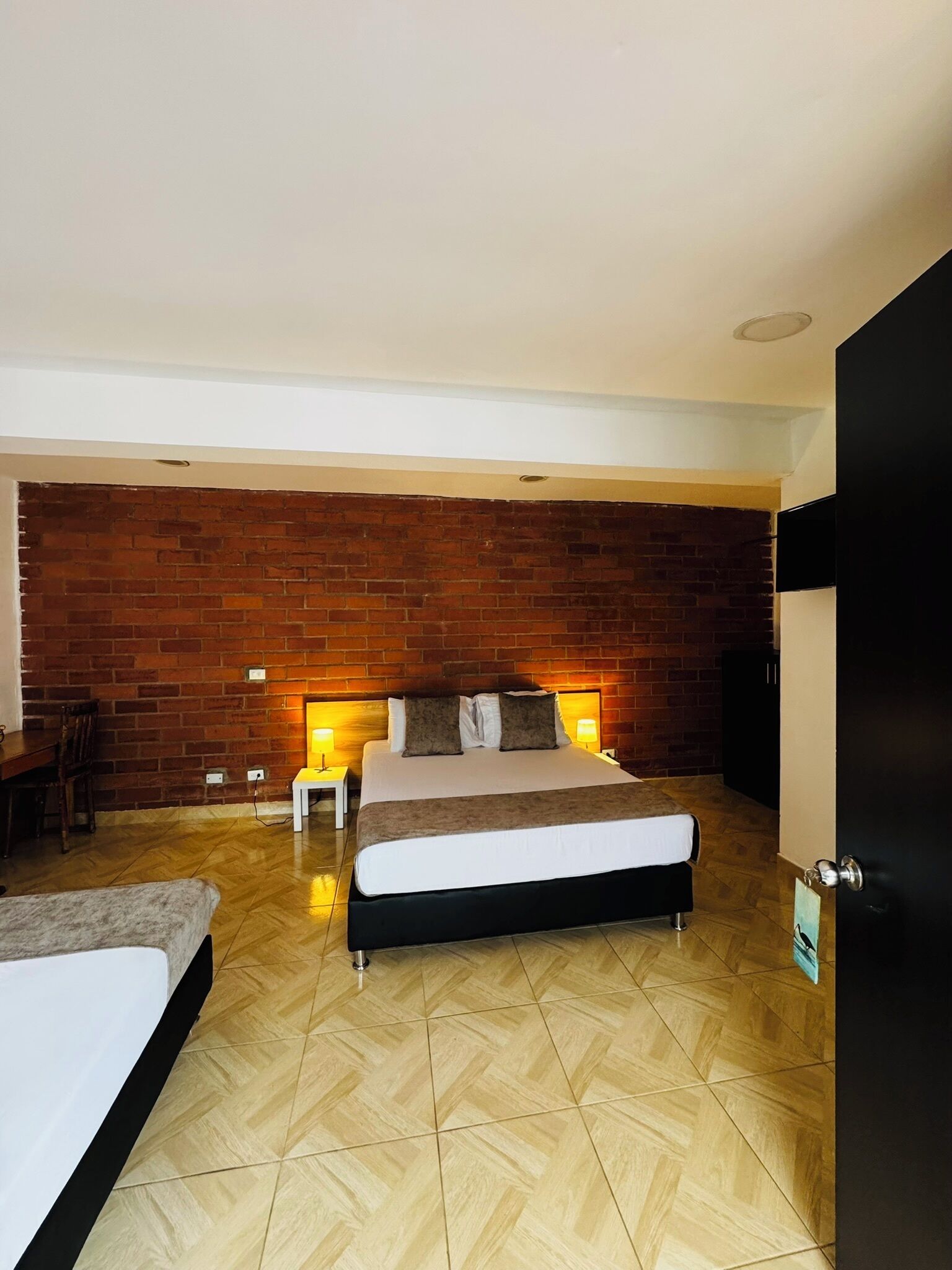 Quarto de hotel com parede de tijolos, cama com estrutura preta, pequena mesa com abajur e TV fixada na parede.