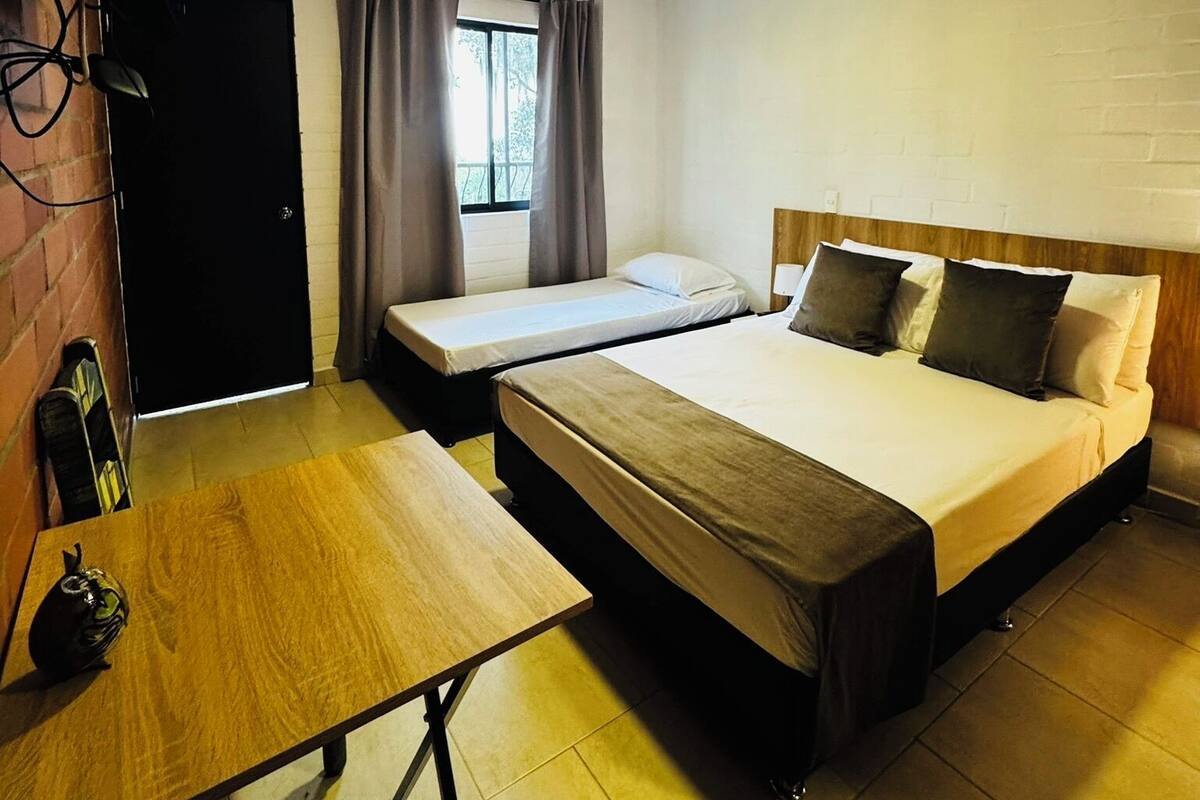 A hotel room with two beds, a desk, a window with curtains, and a brick wall.