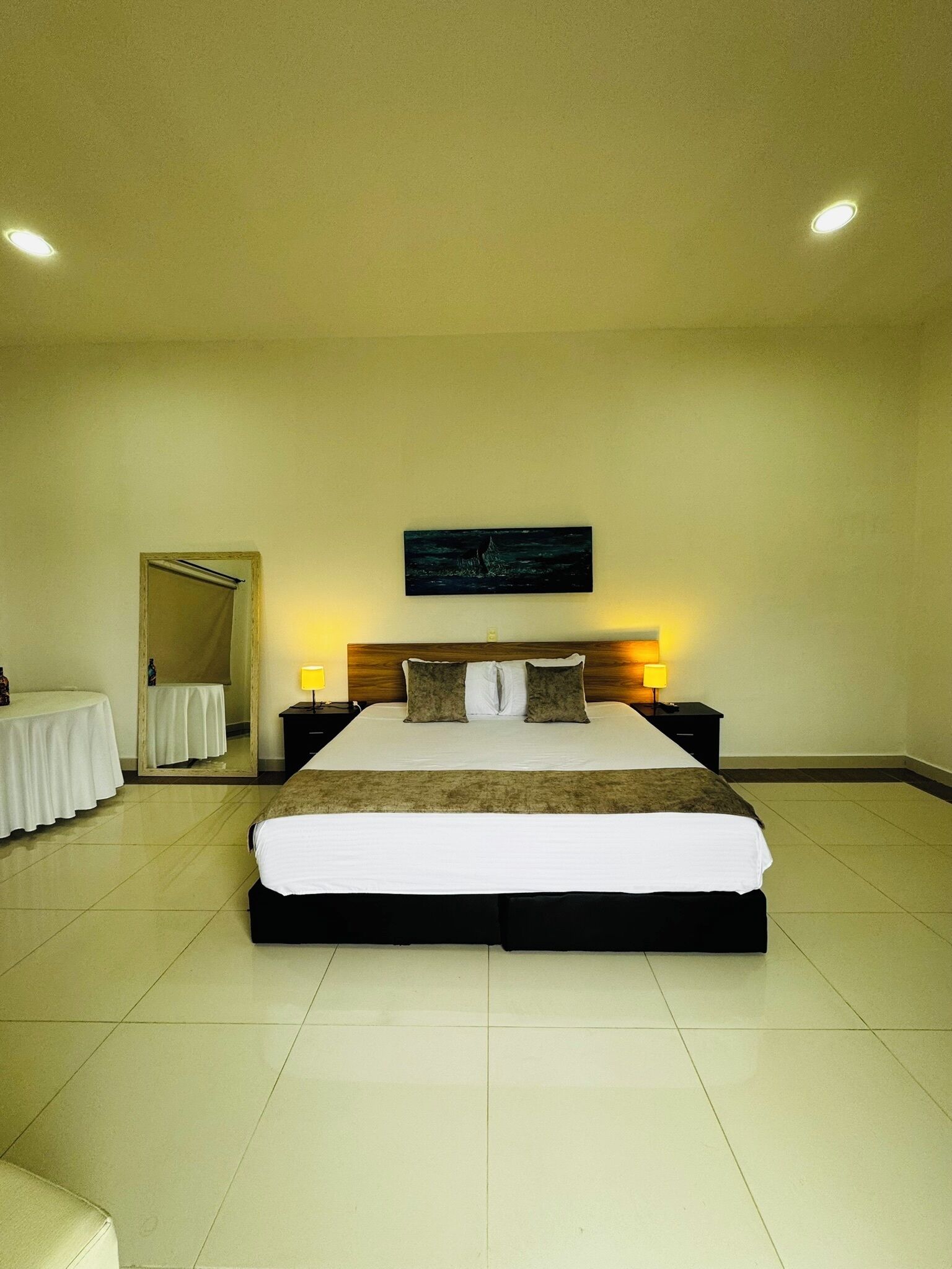 A hotel room with a large bed, a bedside table, a nightstand, a mirror, and a painting on the wall.