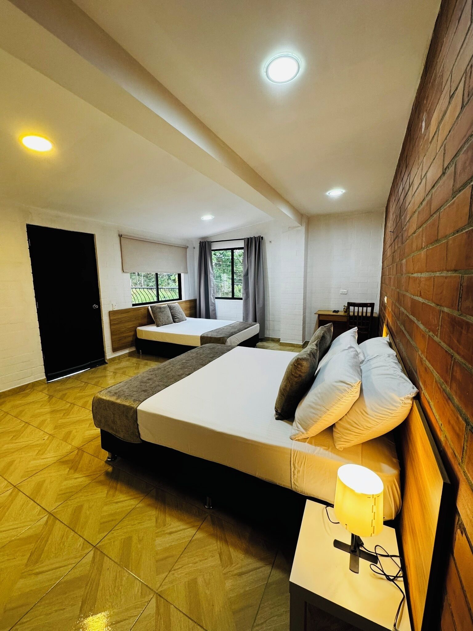 Ein Hotelzimmer mit einem großen Bett, einem Nachttisch mit Lampe, einem Fenster mit Vorhängen und Blick auf Grünflächen.