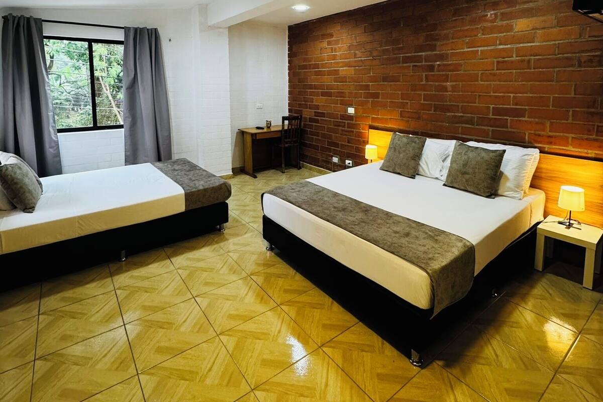 A hotel room with two beds, a brick accent wall, a desk, and a window with curtains.