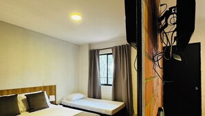 Ein Hotelzimmer mit einem großen Bett, einem kleineren Bett, einem Schreibtisch, einem Stuhl, einem Fernseher und einem Fenster mit Vorhängen.