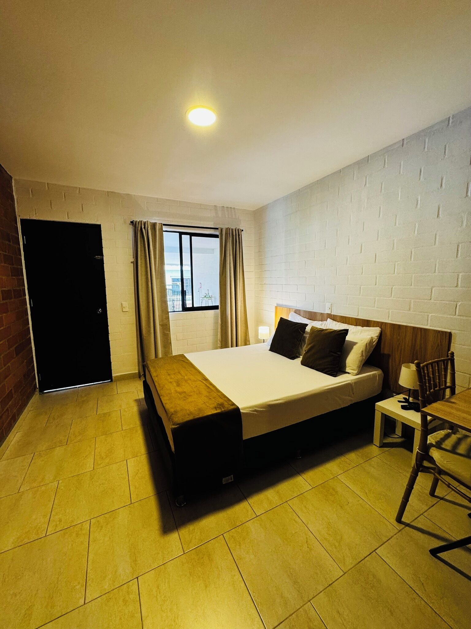 Habitación de hotel con cama, una mesita, una silla, ventana con cortinas y pared de ladrillo.