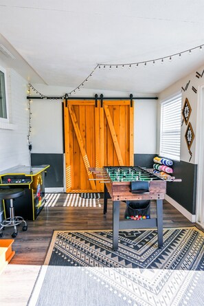 Een kamer met een tafelvoetbaltafel, een bureau met een computer, een stoel en een kamerbreed tapijt met een patroon.
