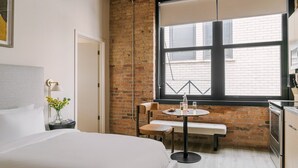 A hotel room with a large bed, a small dining table with chairs, a brick wall, and a window with blinds. - LIVN - West Loop Chicago (Chicago)