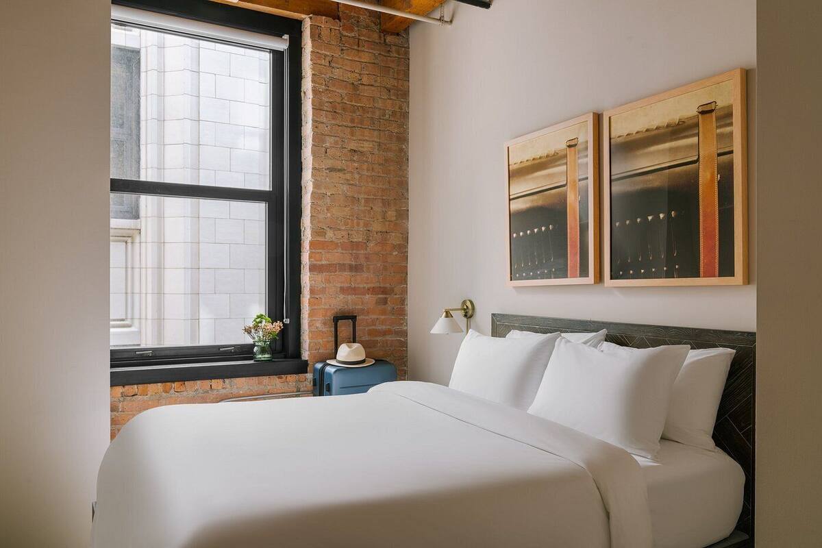 Hotellihuone, jossa on suuri sänky, ikkuna, josta on näkymä rakennuksiin, yöpöytä lampun kanssa ja kaksi kehystettyä kuvaa seinällä.