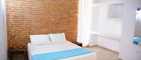 A room with a brick wall, a bed with white bedding, a blue blanket, a small wooden nightstand, a white air conditioning unit, and a white shelf with a mirror.