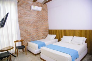 Habitación de hotel con dos camas individuales, una silla de madera, una mesita pequeña, un televisor montado en la pared y una pared de ladrillo.