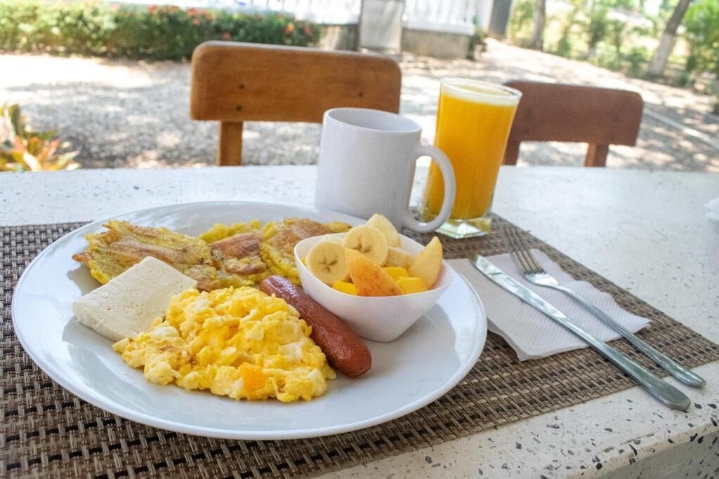 Un plato de desayuno con huevos revueltos, salchicha, tostadas y un bol de fruta, acompañado de un vaso de jugo de naranja y cubiertos sobre una mesa al aire libre.