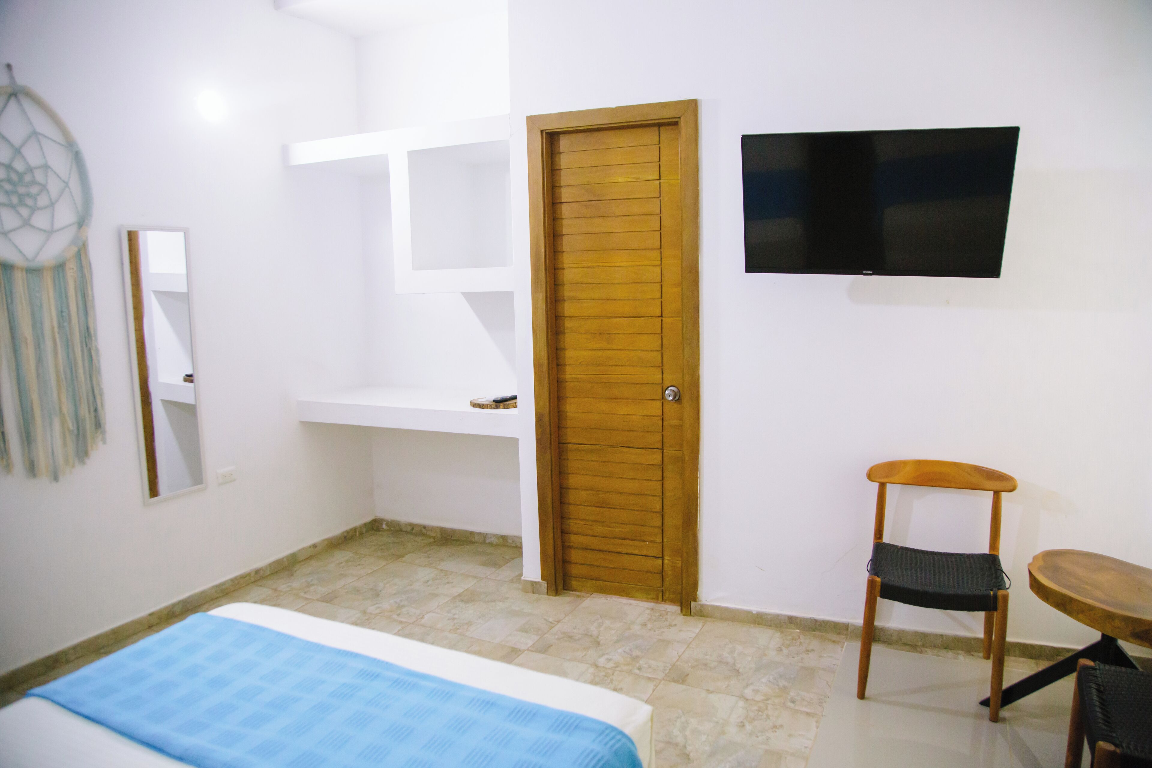A bedroom with a bed, a wooden door, a TV mounted on the wall, a chair, and a small table.