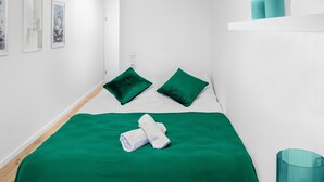 Une chambre d’hôtel avec une couvre-lit verte, deux coussins verts, un mur blanc orné de tableaux encadrés et un vase turquoise posé sur une étagère.