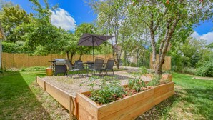 Un patio trasero con camas elevadas para jardín, una parrilla, mobiliario exterior y una sombrilla.