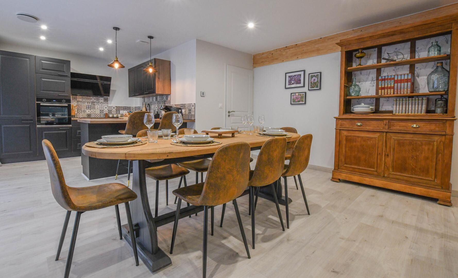 Una cocina moderna con armarios oscuros, una mesa de comedor de madera preparada para seis personas, y un aparador de madera con cristalería.