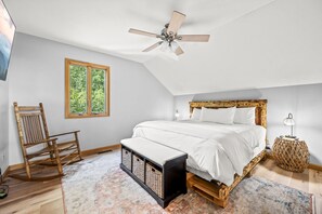 Ein Schlafzimmer mit schrägen Wänden, einem Bett mit Holz-Kopfteil, einem Schaukelstuhl, einem kleinen Tisch und einem Fenster mit Blick auf Grünflächen.