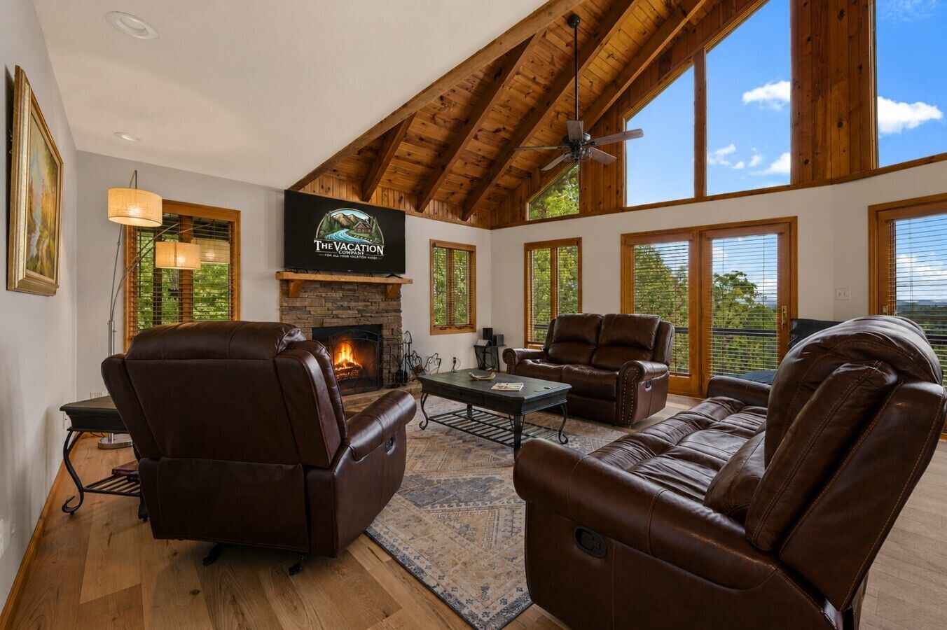 A spacious living room with leather sofas, a fireplace, a television, and large windows offering a view of the outdoors.