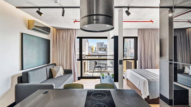 A modern hotel room with a sofa, dining table, and a large window offering a city view. - 1811 Mercure (São Paulo)
