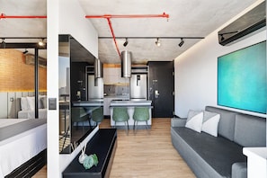 A modern living room with a grey sofa, a coffee table, and a TV. There is a kitchen area with a dining table and chairs. - 1811 Mercure (São Paulo)
