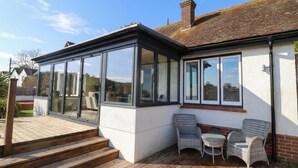 A modern house with a large glass conservatory, a wooden deck, and outdoor seating. - Devona (Seaton)