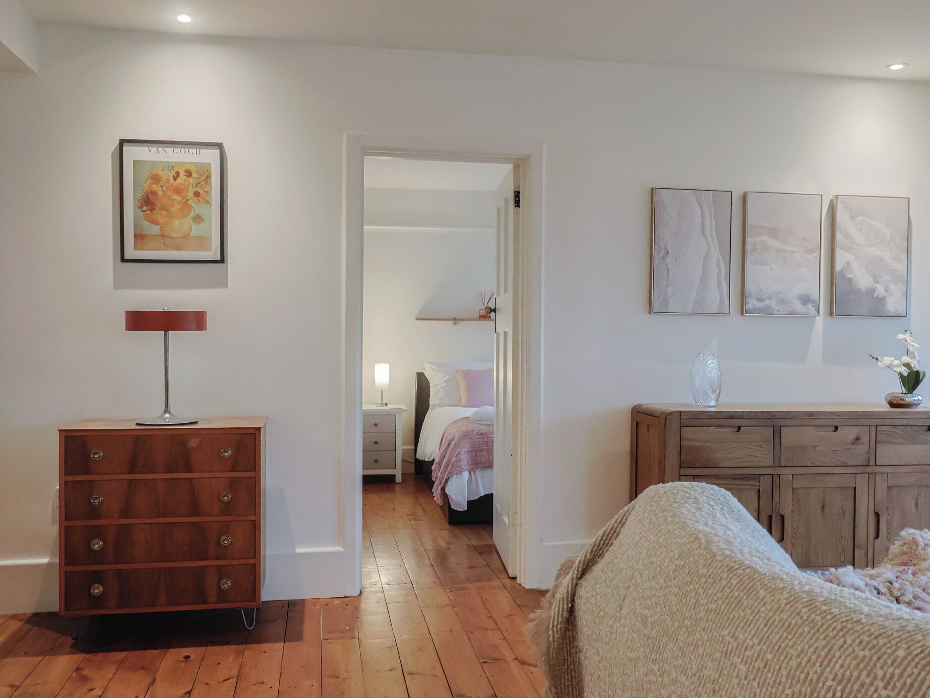 A bedroom with a wooden dresser, a bed with a white blanket, and a nightstand with a lamp. There is a framed artwork on the wall and a door leading to another room.