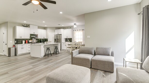Un salon moderne avec un canapé et un pouf, une cuisine avec des meubles blancs et un ventilateur de plafond.