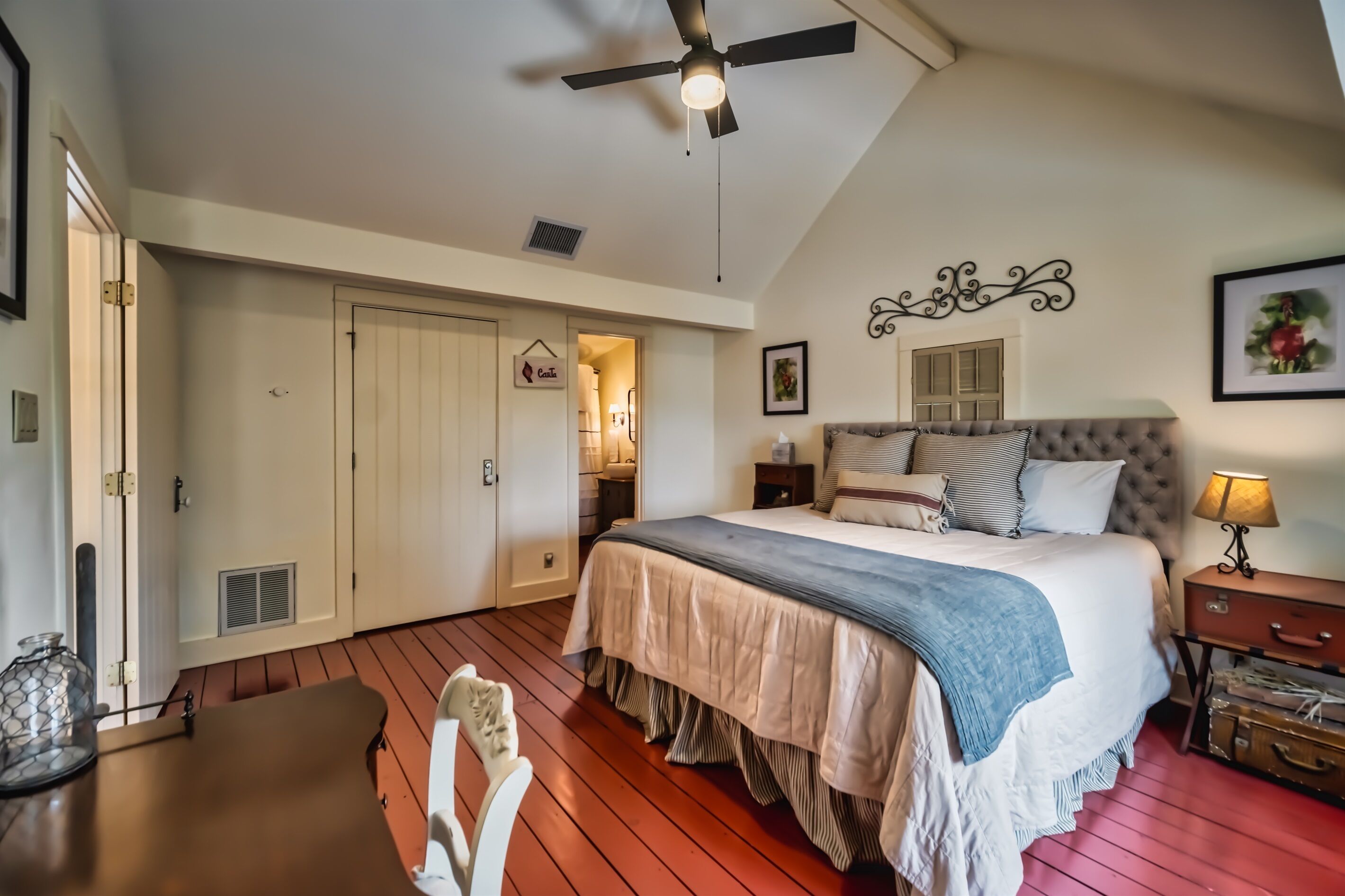 A bedroom with a bed, a ceiling fan, a nightstand, a lamp, a dresser, and a door leading to another room.