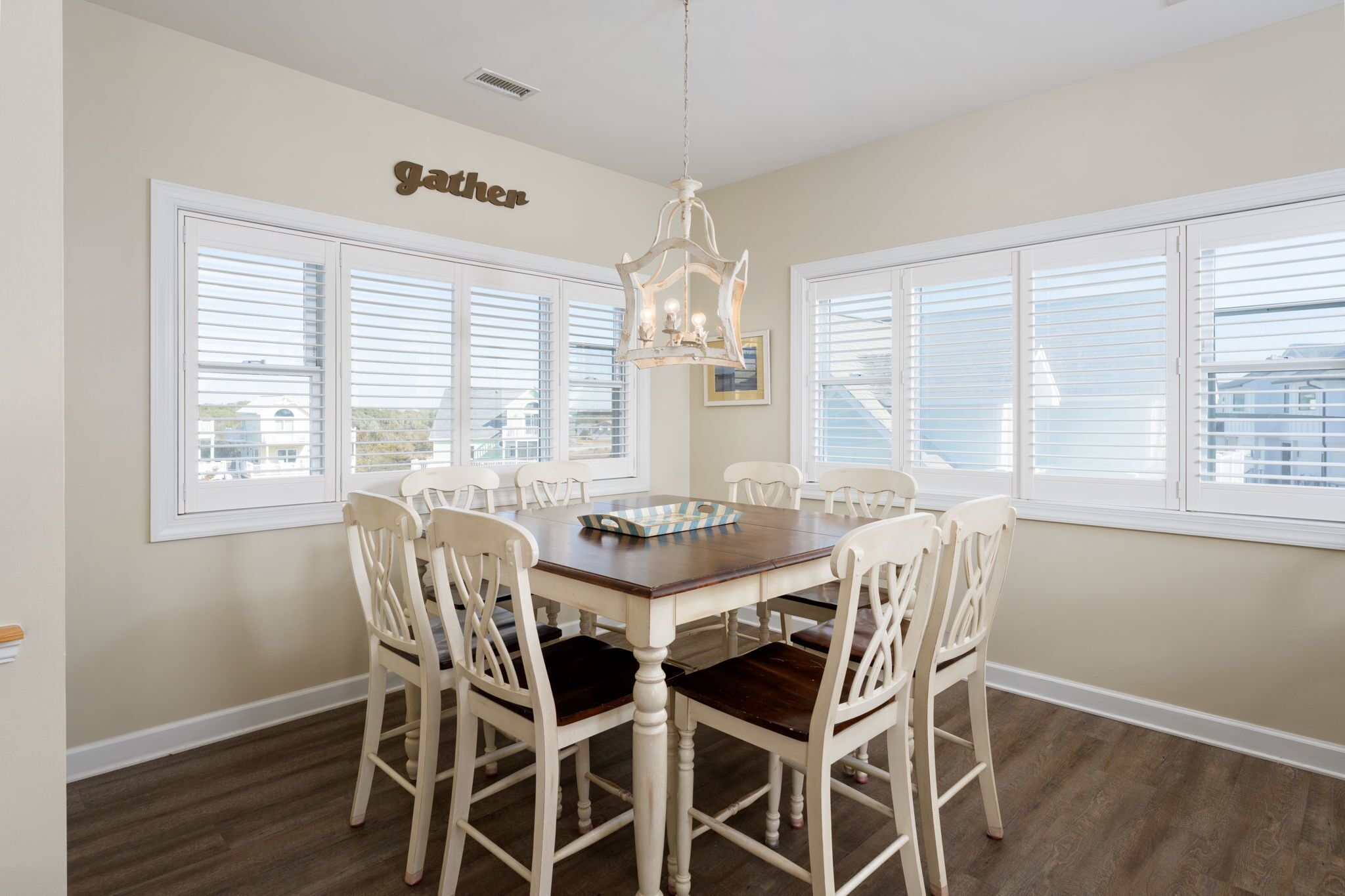 A dining room with a wooden table and white chairs, large windows with blinds, and a chandelier.