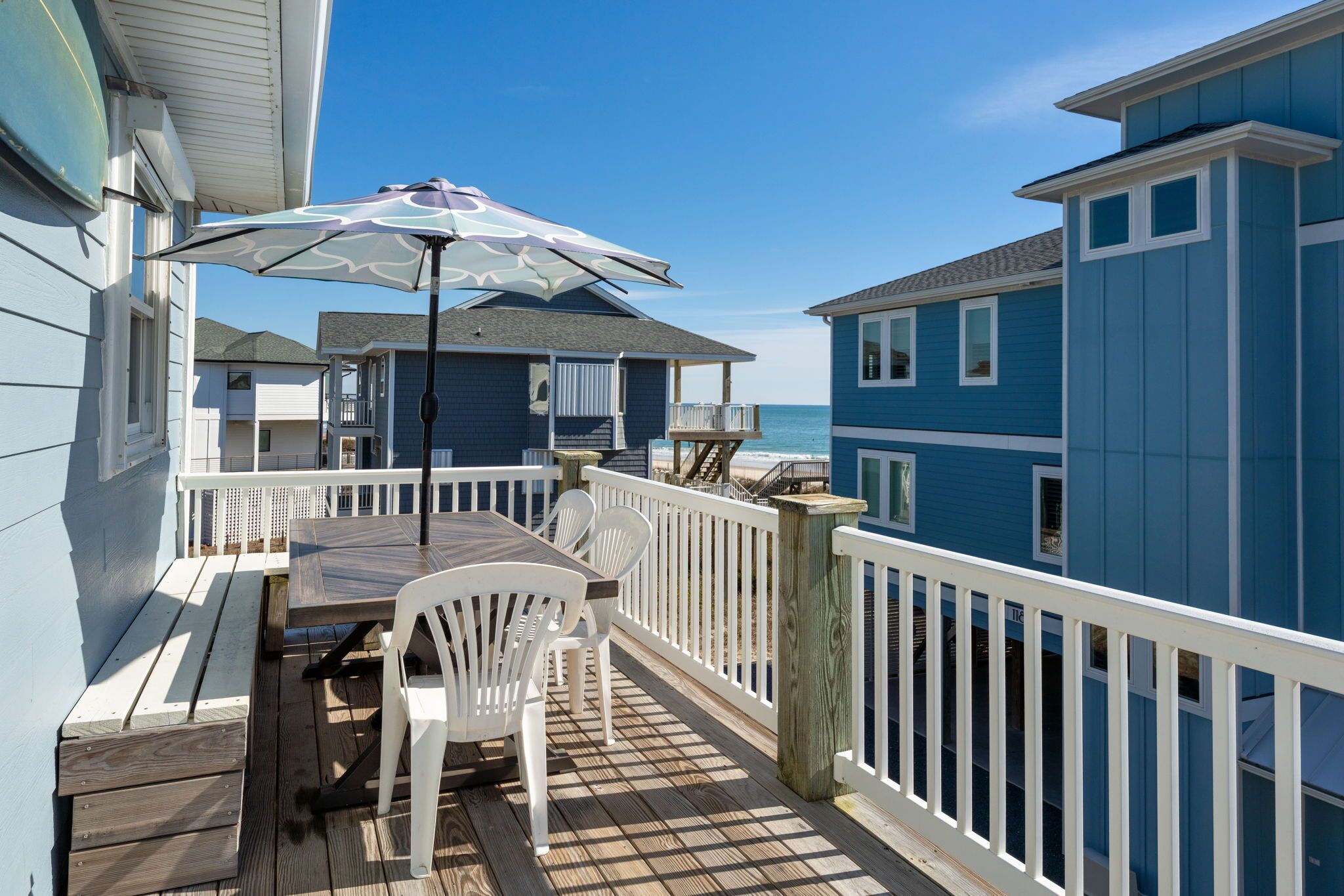 A wooden deck with a table and chairs, a white railing, and a view of the ocean.