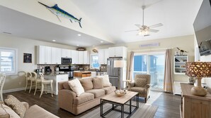 A spacious living room with a large sofa, a coffee table, and a dining area with a kitchen in the background. - Oceanfront home w/ Backyard Beach Access! (North Topsail Beach)