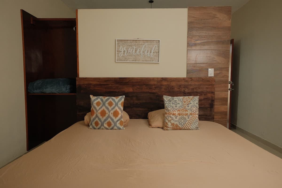 A bedroom with a wooden headboard, a bed with two patterned pillows, a wall-mounted artwork with the word "grateful," and a closet with a blue blanket.