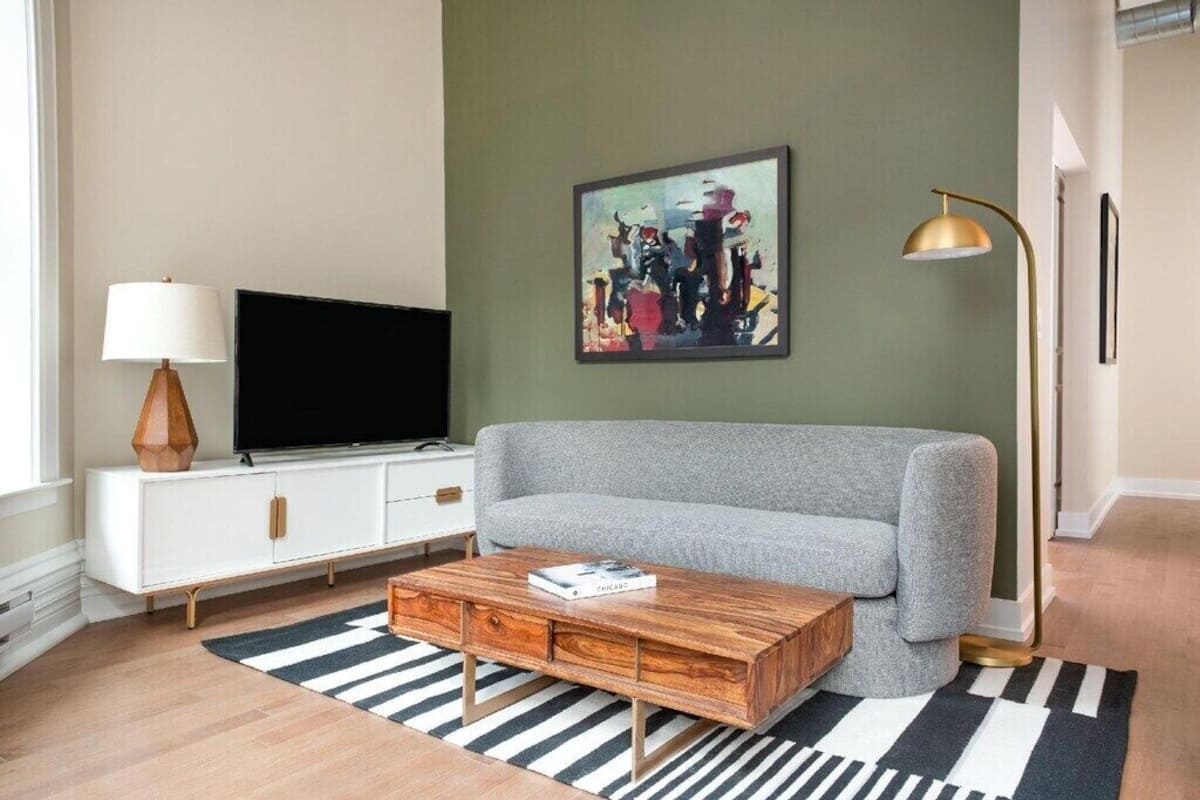 Ett vardagsrum med en grå soffa, ett soffbord i trä, en platt-TV på ett vitt skåp, en tavla på väggen och en golvlampa.